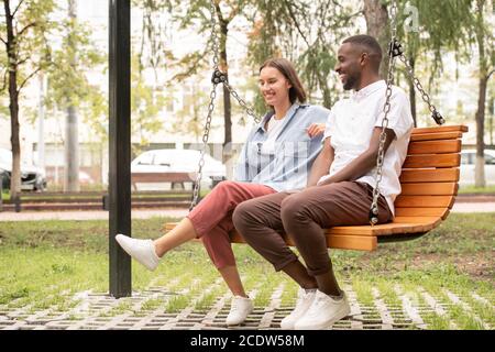 Joyeux jeune couple interculturel en vêtements décontractés assis sur une balançoire en bois dans le parc Banque D'Images