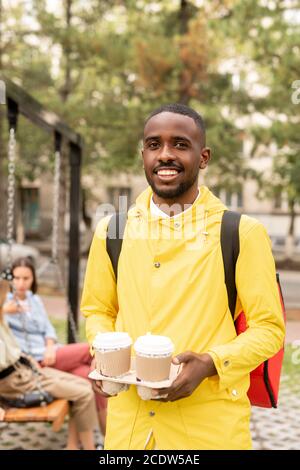 Joyeux jeune homme contemporain courier dans une veste jaune tenant deux boissons Banque D'Images