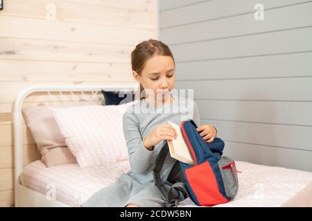 Grave mignonne écolière en robe grise mettant pile de livres dans un sac à dos Banque D'Images