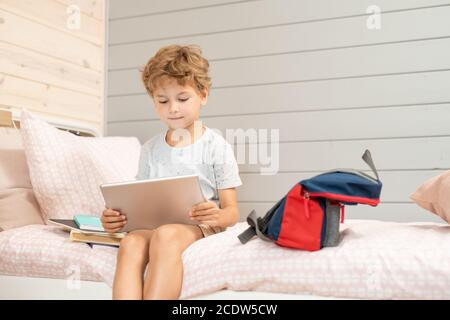 Petit garçon blond mignon avec tablette assis sur le lit et regarder des vidéos en ligne Banque D'Images