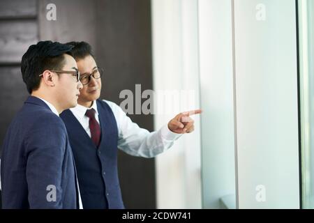 deux dirigeants d'entreprises asiatiques debout sous les fenêtres qui parlent au bureau Banque D'Images