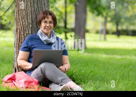 Femme âgée souriante travaillant à distance sur un ordinateur portable dans le parc de la ville. Concept de travail à distance. Banque D'Images