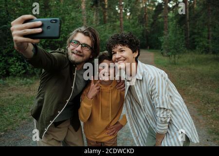 Famille contemporaine de jeunes parents heureux et fils faisant le selfie à l'extérieur Banque D'Images