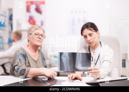 Médecin discutant avec la patiente âgée de sa blessure à la jambe tout en tenant des rayons X dans le bureau de l'hôpital. Medic portant une couche blanche travaillant sur l'ordinateur. Banque D'Images