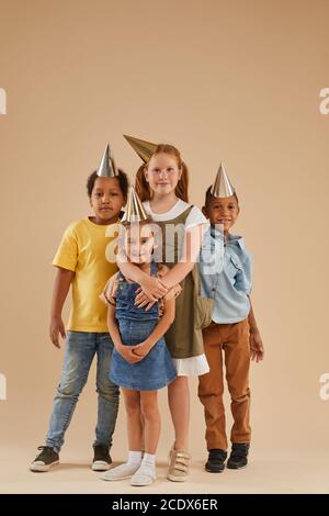 Portrait vertical sur toute la longueur de divers groupes d'enfants portant chapeaux de fête regardant l'appareil photo tout en se tenant contre un fond uni en studio Banque D'Images