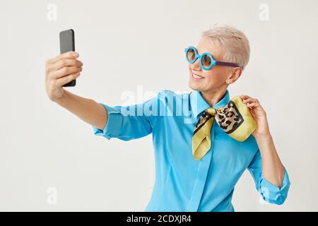 Femme élégante senior en lunettes de soleil se posant à l'appareil photo sur elle téléphone portable sur fond blanc Banque D'Images