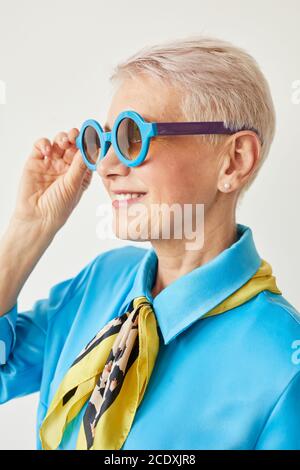 Élégante femme âgée avec cheveux courts blonds portant des lunettes de soleil et souriant isolé sur fond blanc Banque D'Images