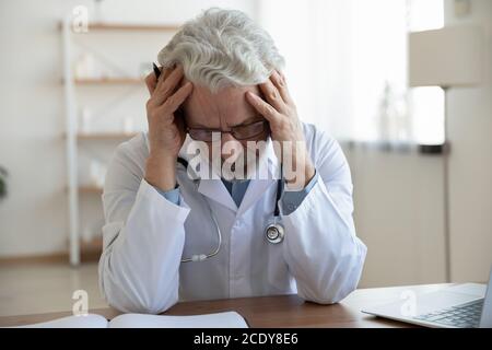 Épuisé malheureux mature médecin portant des lunettes assis au bureau Banque D'Images