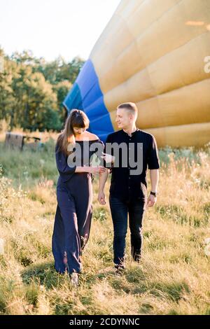 Photo d'été en plein air d'un jeune couple joyeux marchant au coucher du soleil dans un champ vert, posant à la caméra devant un ballon d'air chaud jaune, se préparant pour Banque D'Images