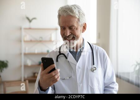 Sourire un médecin mûr portant un uniforme blanc regardant l'écran du smartphone Banque D'Images