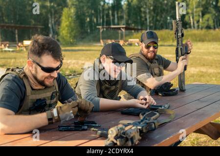 Jeunes hommes positifs dans les brassards de corps assis à une table en bois et discuter tout en rechargeant des armes Banque D'Images