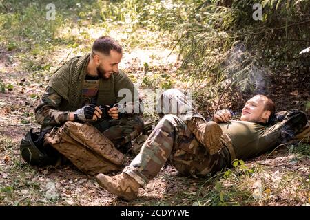 De jeunes soldats positifs en uniforme militaire fument de la cigarette et bavardent tout en se reposant dans un abri forestier Banque D'Images