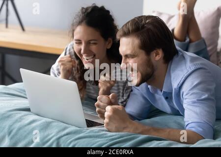 Un jeune couple surjoyeux allongé sur le lit, regardant l'écran d'un ordinateur portable. Banque D'Images