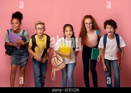 Enfants multiethniques stupides avec des fournitures scolaires qui font des visages amusants fond rose Banque D'Images