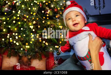 Un petit garçon joyeux devant l'arbre de Noël entre les mains des mères Banque D'Images