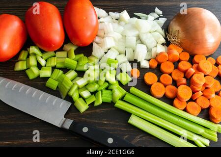 Hacher des légumes sur une planche à découper en bois foncé : céleri, oignon et carottes hachés avec des tomates prune Banque D'Images