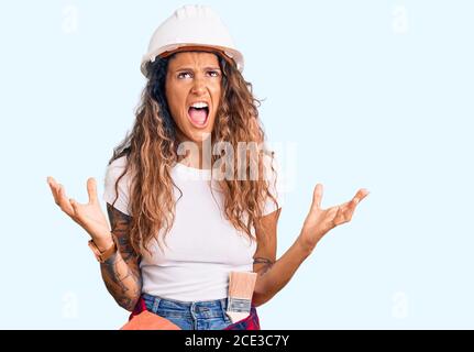 Jeune femme hispanique avec tatouage portant un casque et des vêtements de constructeur fou et fou criant et hurlant avec l'expression agressive et les bras levés. fr Banque D'Images