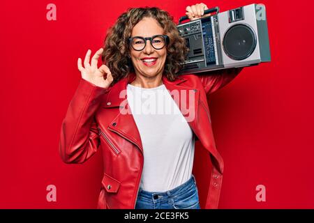Moyen âge brunette hipster femme tenant la boombox de musique rétro sur fond rouge isolé faisant signe ok avec les doigts, souriant gestining exce Banque D'Images
