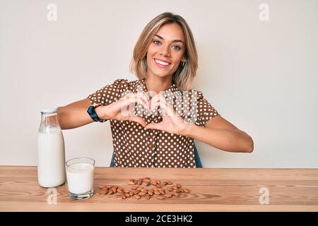 Belle femme caucasienne boit du lait d'amande sain souriant en amour faisant le symbole de forme de coeur avec les mains. Concept romantique. Banque D'Images