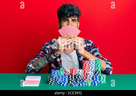 Jeune homme hispanique jouant au poker de jeu couvrant le visage avec des cartes souriant avec un sourire heureux et cool sur le visage. Montrant des dents. Banque D'Images