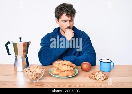 Jeune caucasien avec des cheveux bouclés assis sur la table ayant le petit déjeuner se sentir mal et la toux comme symptôme de rhume ou de bronchite. Soins de santé c Banque D'Images