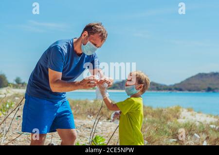 Volontaires dans le masque médical bleu. Père met des gants sur son fils pour ramasser les ordures qui polluent la plage près de la mer. Problème de déchets déversés Banque D'Images