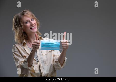 Tête portrait belle blonde femme regardant l'appareil photo tenir médical ou masque chirurgical de couleur bleue Banque D'Images