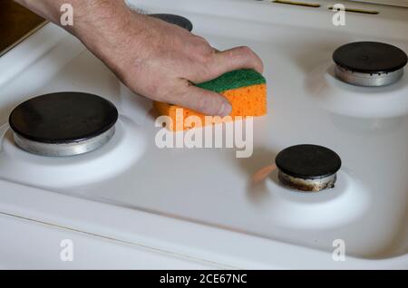 L'homme nettoie la cuisinière avec une éponge de cuisine. Main masculine avec une éponge de cuisine orange. Homme d'âge moyen, caucasien. Service de nettoyage. Mise au point sélective. Banque D'Images