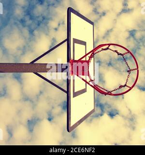 Vieux panneau arrière de basket-ball négligé avec un cerceau rouillé au-dessus du court de rue. Ciel bleu ciel nuageux dans le fond de bckground. Filtre rétro Banque D'Images