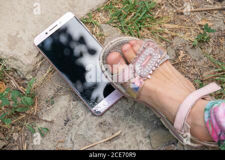 pied d'enfant dans des sandales marches sur le téléphone avec un écran cassé reposant sur le sol Banque D'Images