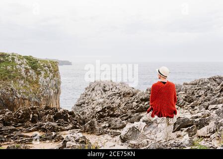 Vue arrière d'une femme imreconnaissable dans des vêtements élégants, assise au bord de la falaise rocheuse contre un ciel nuageux et admirant le paysage marin tout en profitant de vacances sur la côte asturienne de l'Espagne Banque D'Images
