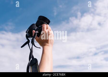 Rognez un photographe méconnu dans des vêtements élégants debout avec le bras relevé prise en main d'un appareil photo professionnel dans un ciel bleu nuageux Banque D'Images