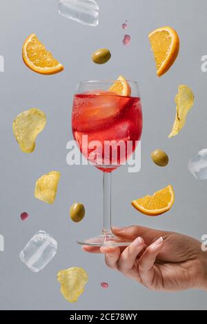 Crop femelle avec un verre d'alcool rouge cocktail garni tranches d'orange et olives vertes au-dessus du sol sur fond gris Banque D'Images