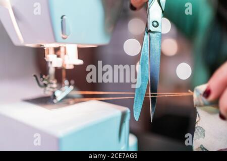 Récolte d'un artisan méconnaissable qui prolonge le fil fin tout en coupant avec des ciseaux sur une machine à coudre métallique moderne sur une table en atelier Banque D'Images