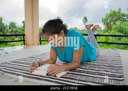 Une femme en haut bleu lit un roman sur un jour couvert dans un environnement d'arbres en arrière-plan. Elle repose sur un tapis, ses cheveux attachés dans un petit pain. Banque D'Images