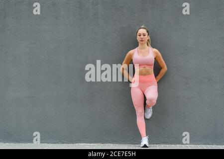 Corps complet de l'athlète féminine en tenue de sport et baskets debout sur une jambe sur un remblai presque gris mur et regarder la caméra Banque D'Images