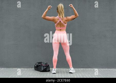 Vue arrière d'une sportive blonde fine et non reconnaissable, en tenue active, qui fait la démonstration de biceps tout en se tenant près d'un sac de sport sur la chaussée, sur fond gris Banque D'Images