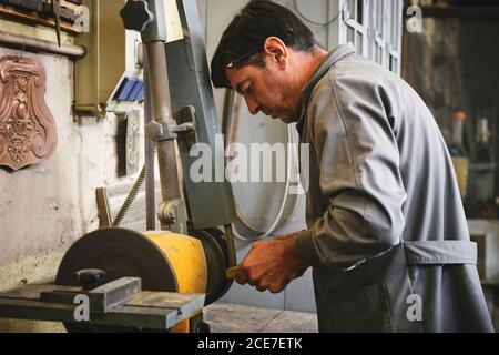 Vue latérale d'un bijoutier homme mature professionnel pour le meulage de vêtements de travail barre métallique sur la machine à polir pendant les travaux en atelier Banque D'Images