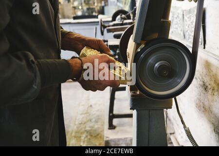 Vue latérale de la récolte de l'orfèvre mâle non reconnaissable sur les bords de polissage détail métallique lors de l'utilisation d'une meuleuse électrique en atelier Banque D'Images