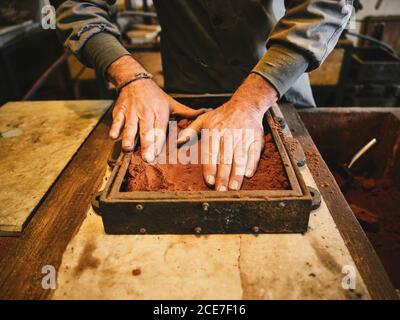 Rognez du sable de pressage d'orfèvre mâle non reconnaissable dans la boîte de moulage avec mains lors de la préparation du moule pour le boîtier métallique en atelier Banque D'Images