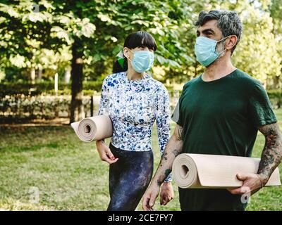 Homme positif adulte tatoué et femme gaie dans des vêtements décontractés et des masques de protection avec tapis de yoga roulés parlant tout en marchant dans le parc vert après avoir pratiqué le yoga ensemble Banque D'Images