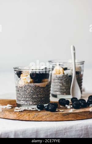 Bocaux en verre avec de délicieux puddings au chia faits de frais savoureux lait d'amande et graines de chia au miel recouvert de mûres et une banane en tranches servie sur un panneau de bois sur fond blanc Banque D'Images