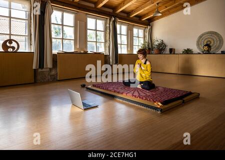 Femme calme dans les vêtements de sport assis sur un tapis dans un studio spacieux Et pratiquer le yoga à Padmasana tout en filmant un tutoriel en ligne sur ordinateur portable Banque D'Images