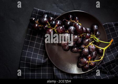 Vue de dessus d'un bouquet de délicieux raisins rouges mûrs dans un bol placé sur une serviette à carreaux sur une surface noire Banque D'Images