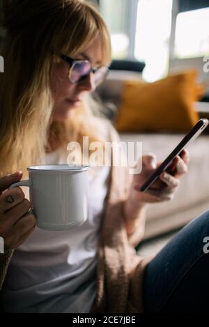 Contenu femme assis sur le sol sur la moquette douce et de prendre photo sur téléphone portable tout en buvant une boisson chaude dans un mug relaxant à la maison Banque D'Images