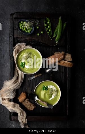 De dessus vue de deux bols de crème de pois placés sur un plateau en bois et une surface sombre, servi avec du pain et décoré avec des pois entiers et de la crème aigre Banque D'Images