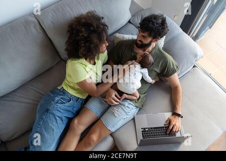 Par-dessus la mère noire et le bébé mignon couché Canapé avec un père arabe travaillant sur un ordinateur portable à la maison Banque D'Images