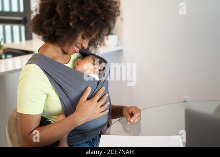 Joyeuse femme afro-américaine assise à la table à la maison avec bébé endormi en toile Banque D'Images