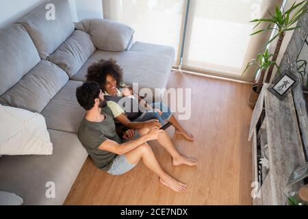 Du contenu ci-dessus, mère et père multiracial jouant avec adorable bébé en se reposant sur le sol dans un appartement moderne Banque D'Images