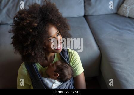 D'en haut douce mère afro-américaine assis sur le sol avec adorable bébé dormant en toile et regardant l'appareil photo Banque D'Images